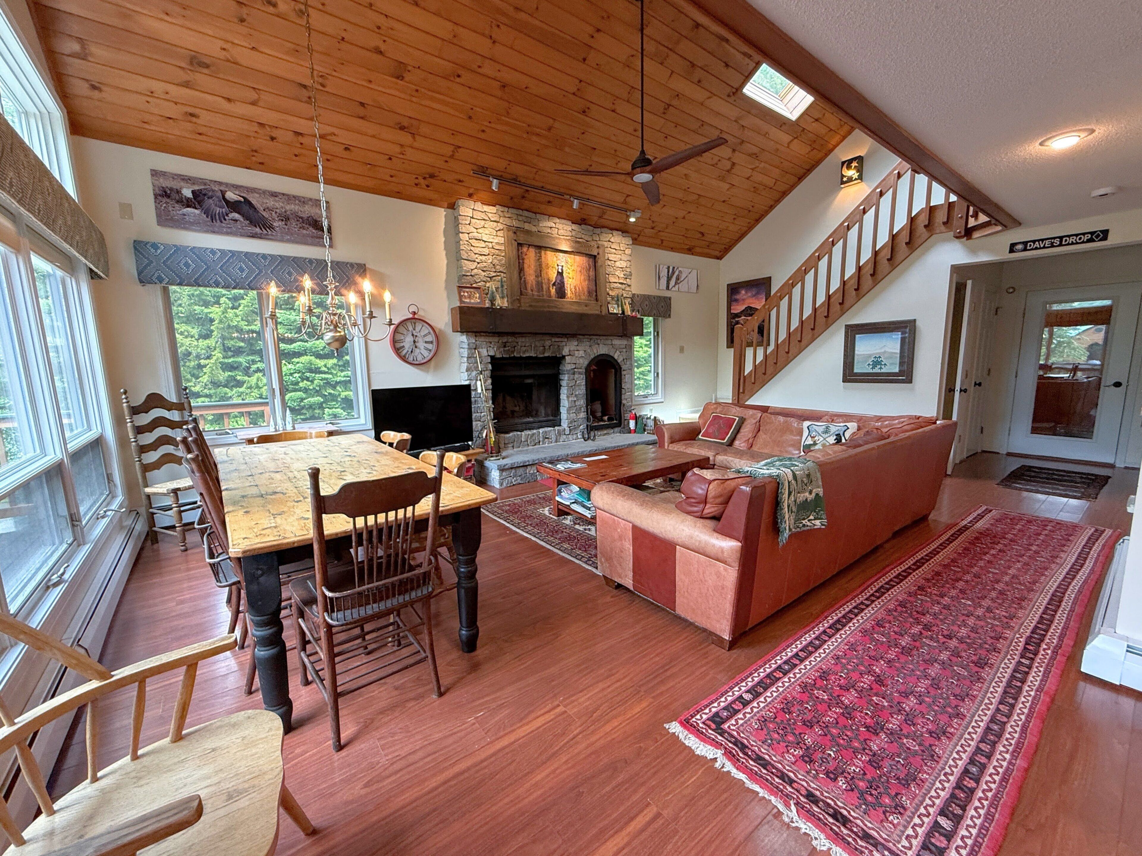 A spacious living area with a fireplace, wooden flooring, a dining table with chairs, a sofa, and a staircase.