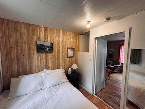 A bedroom with a wooden wall, a bed, a nightstand with a lamp, and a door leading to another room.