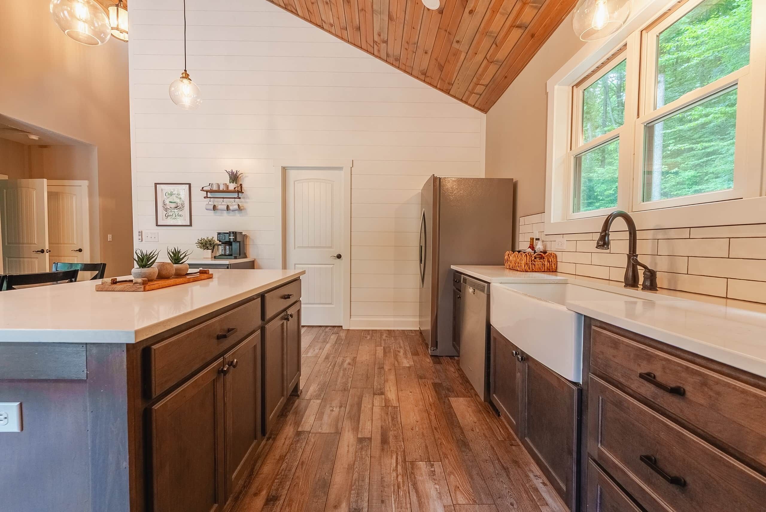 Une cuisine moderne avec un plancher en bois, un îlot central, des armoires intégrées et une fenêtre donnant sur un espace verdoyant.