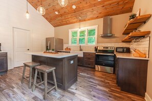 A modern kitchen with a wooden ceiling, a central island with stools, a built-in oven, and a microwave. - Scout's Retreat (Green Township)