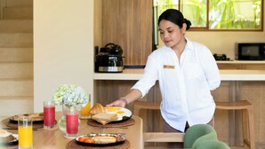 Eine Person serviert Frühstück an einem Holztisch mit verschiedenen Speisen, darunter Toast, Eier und ein Croissant.
