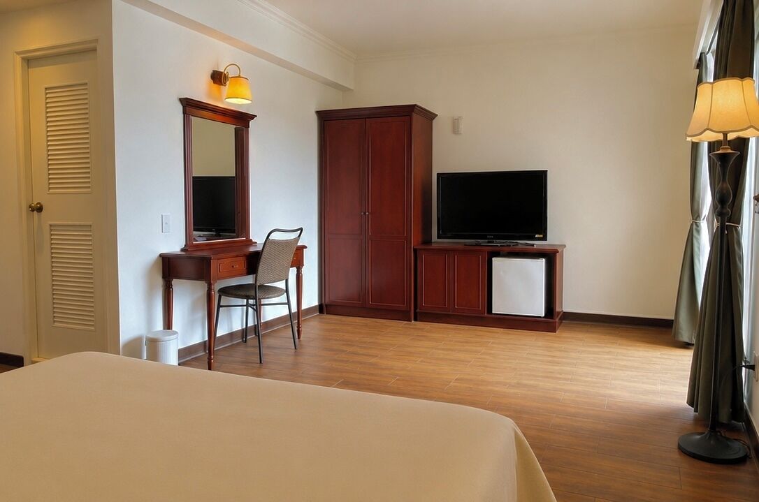 A hotel room with a bed, a desk with a chair, a television, a wardrobe, and a lamp.