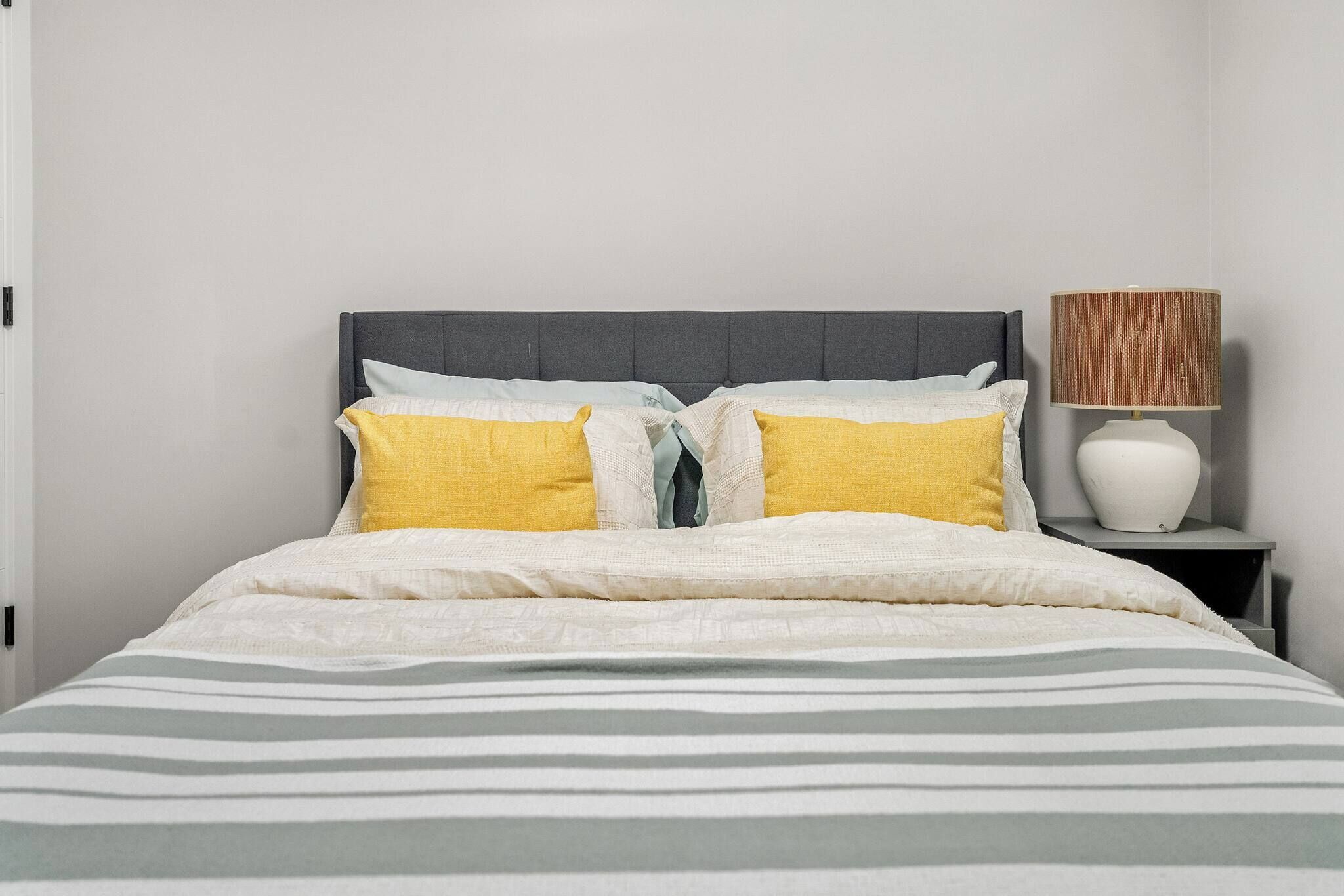 Ein Bett mit grauem Kopfteil, weißen und gelben Kissen, einer gestreiften Decke und einer Nachttischlampe mit gewebtem Lampenschirm.