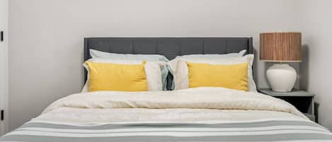 Un lit avec une tête de lit grise, des oreillers blancs et jaunes, une couverture à rayures et une lampe de chevet avec un abat-jour en osier.