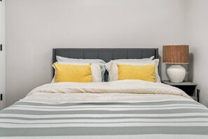 A bed with a grey headboard, white and yellow pillows, a striped blanket, and a bedside lamp with a woven shade. - Latobir Modern,stylish 2BR Suite Private Entrance (Calgary)