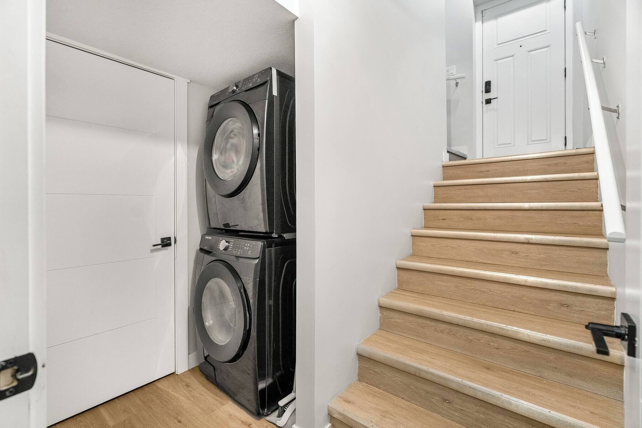 A laundry room with two stacked washers and dryers, a wooden staircase, and a white door.