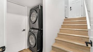 A laundry room with two stacked washers and dryers, a wooden staircase, and a white door.