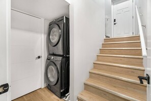 Una sala de bugaderia amb dues piles de rentadores i assecadores, una escala de fusta i una porta blanca.