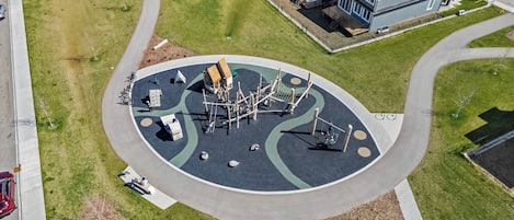 Une vue aérienne d’un quartier résidentiel avec une aire de jeux au centre, entourée de maisons et d’une route sinueuse.