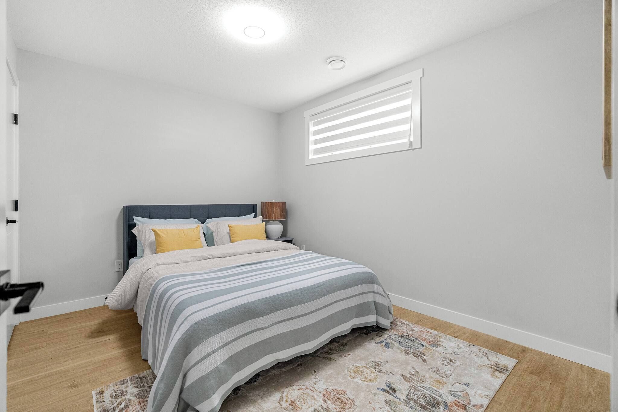 Un dormitorio con cama, mesita de noche, lámpara, ventana con persianas y una alfombra estampada.