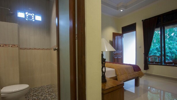 Habitación de hotel con inodoro, cama, mesita de noche de madera y ventana con vistas a zonas verdes.