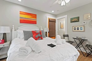 A neatly made bed with white linens, a red and black pillow, and a folded towel. There is a small table with two cups and a chair.