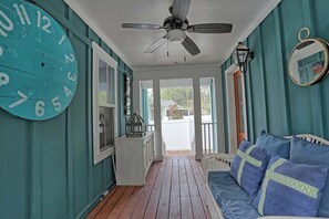 A porch with a ceiling fan, a large clock, a white sofa with patterned cushions, and a view of the outside.
