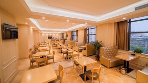 Ein Hotelrestaurant mit mehreren Tischen und Sitzecken, einem an der Wand befestigten Flachbildfernseher und großen Fenstern mit Blick auf die Stadt.