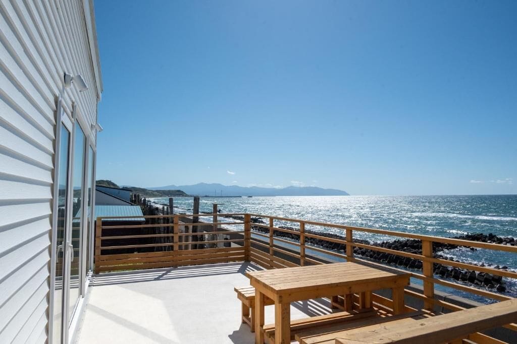 Un balcon avec une table en bois et des bancs, donnant sur une côte rocheuse et l’océan.