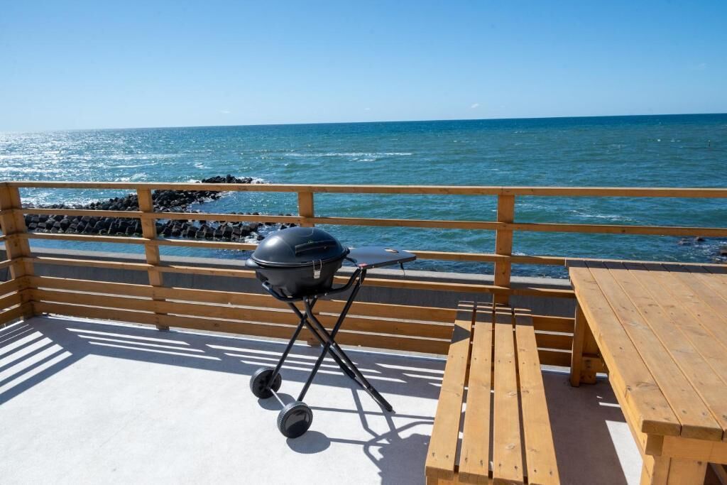 A wooden deck with a barbecue grill and a table, overlooking the ocean.
