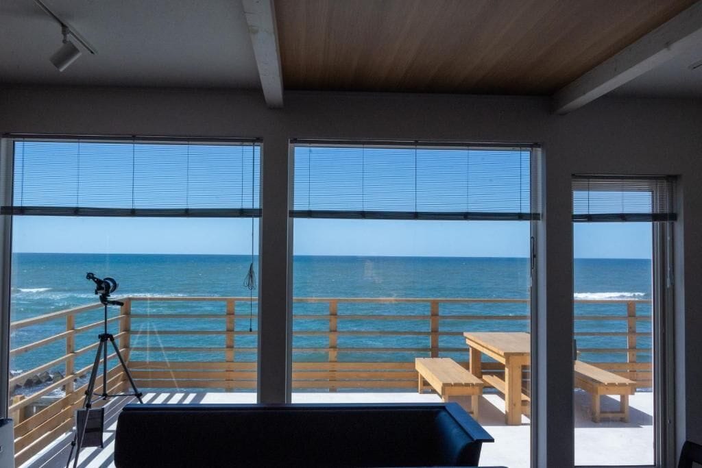 A room with large windows overlooking the ocean, a telescope on the left, and a wooden table and bench on the right.