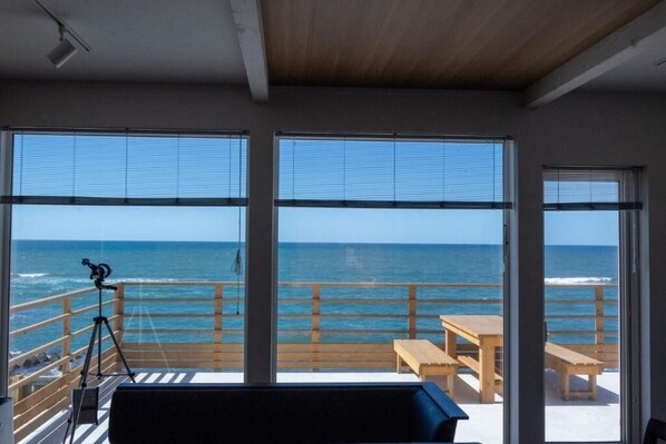 A room with large windows overlooking the ocean, a telescope on the left, and a wooden table and bench on the right.