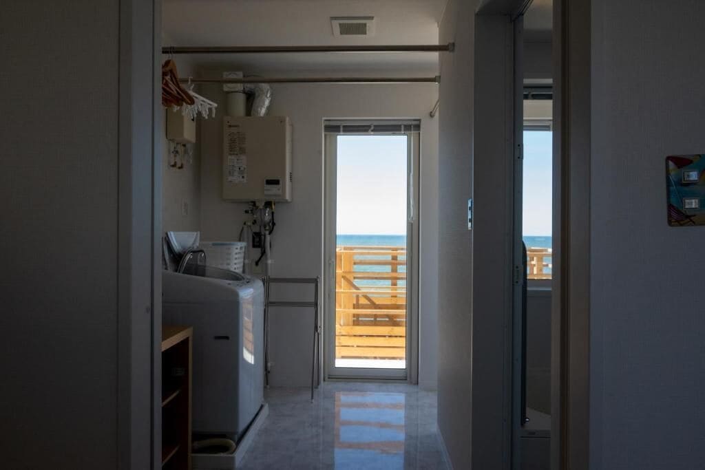 A narrow hallway leading to a balcony with a wooden railing, a small kitchenette with a refrigerator, and a door leading outside.
