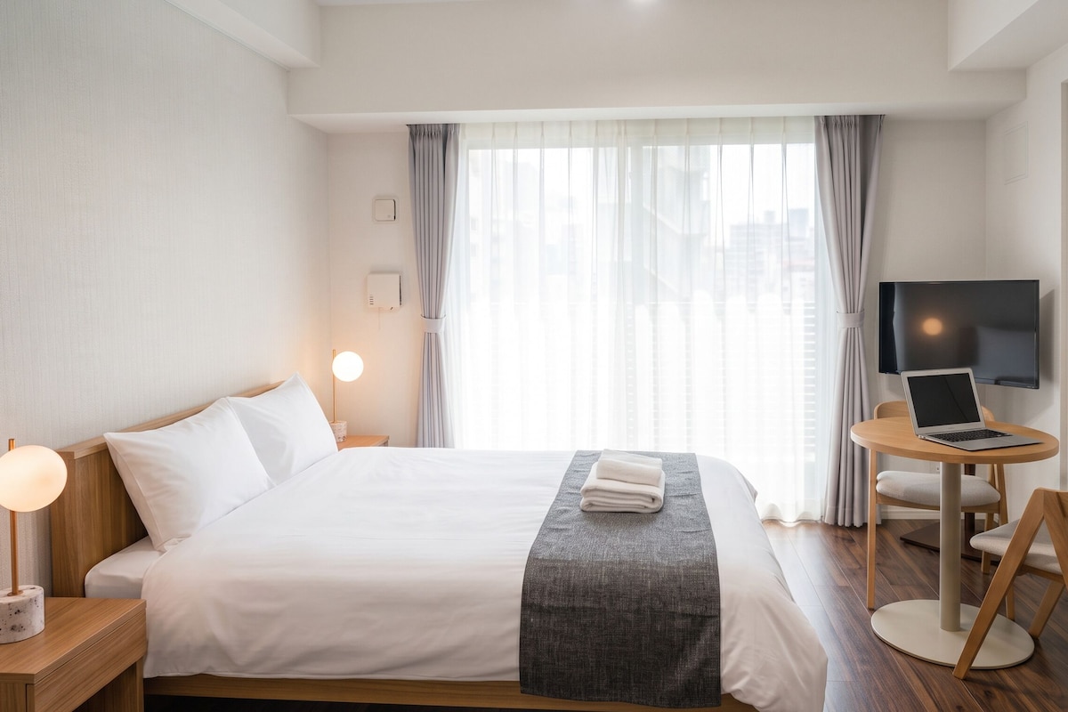 Quarto de hotel com cama, escrivaninha com laptop, televisão e janela com cortinas.
