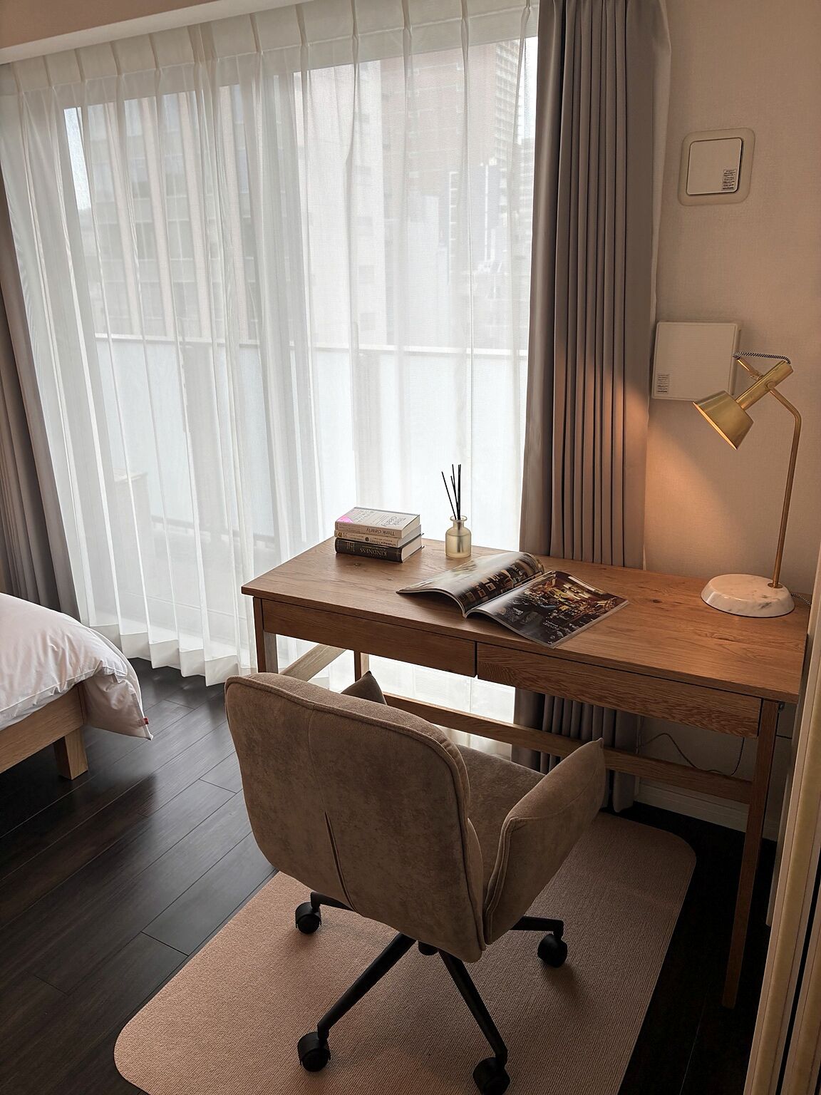 A hotel room with a wooden desk, a chair, a bedside table with a lamp, and a window with sheer curtains.