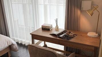 A hotel room with a wooden desk, a chair, a bedside table with a lamp, and a window with sheer curtains.
