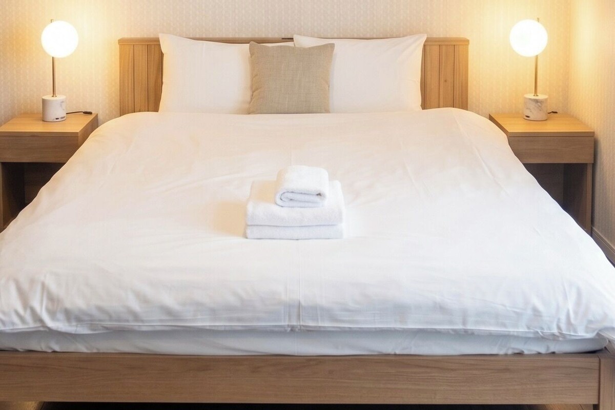 A neatly made bed with white linens and a folded towel on the center. There are two bedside tables with lamps on either side of the bed.