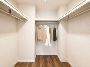 A walk-in closet with a wooden floor, white walls, and a hanging rod with clothes. - Dash Living Toranomon (Minato-ku)