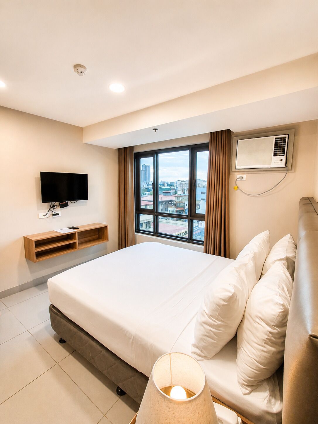 A hotel room with a large bed, a flat-screen TV mounted on the wall, an air conditioning unit, and a view of the cityscape through the window.