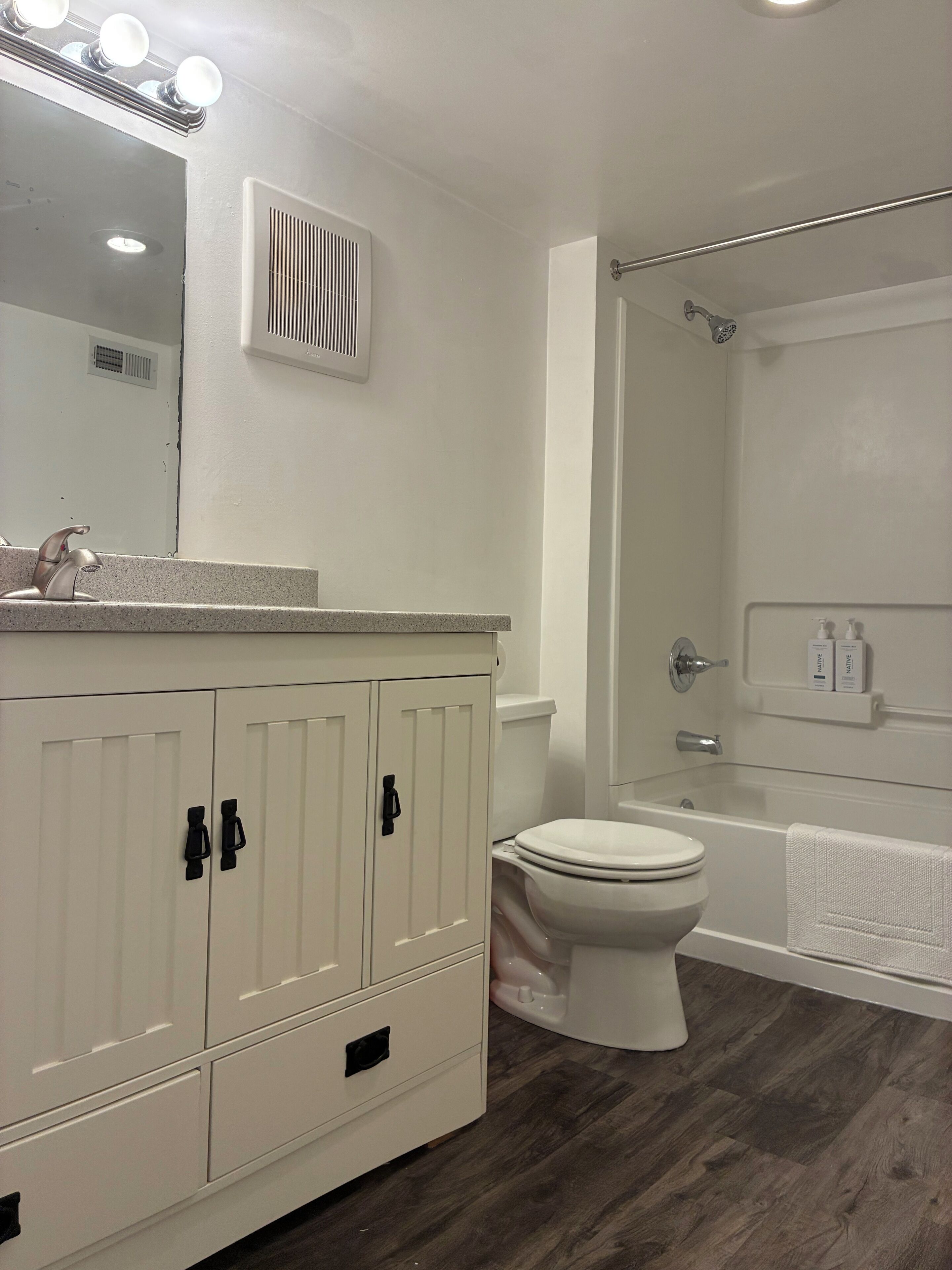 A bathroom with a white vanity, a toilet, a shower area, and a mirror with lights.