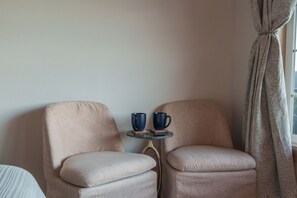Ein Zimmer mit zwei Sesseln, einem kleinen runden Tisch mit zwei blauen Tassen und einem Fenster mit Vorhängen.