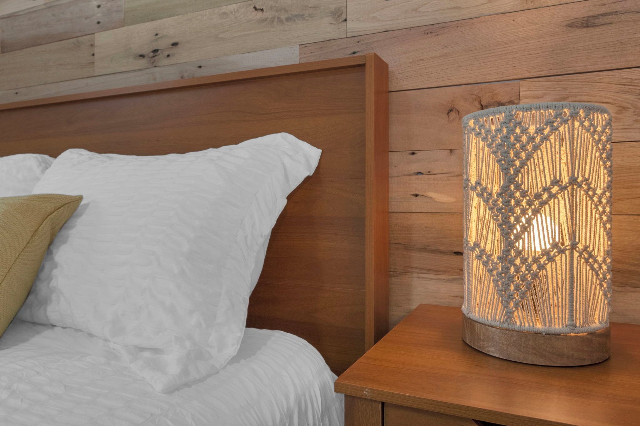 A bed with white bedding and a beige pillow, a wooden nightstand with a macrame lamp, and a wooden headboard.