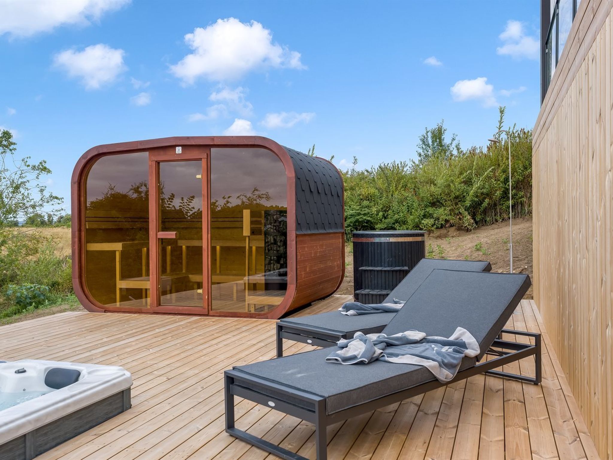 A modern outdoor deck with a glass-enclosed structure, lounge chairs, and a hot tub.