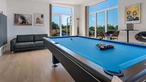 Een modern binnenzwembad met een pooltafel, een bank en grote ramen met uitzicht op de omgeving.