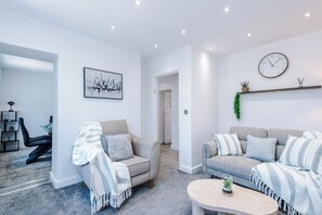 A modern living room with two sofas, a coffee table, a chair, a framed cityscape artwork, a clock, and a shelf with plants. - Modern Chorlton Home | Parking | City and Airport (Manchester)