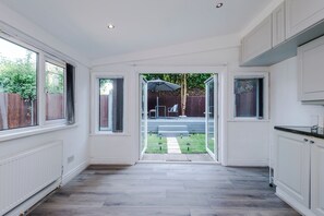 A spacious kitchen with white cabinetry, a large window, and a view of a backyard with a patio and lawn. - Modern Chorlton Home | Parking | City and Airport (Manchester)