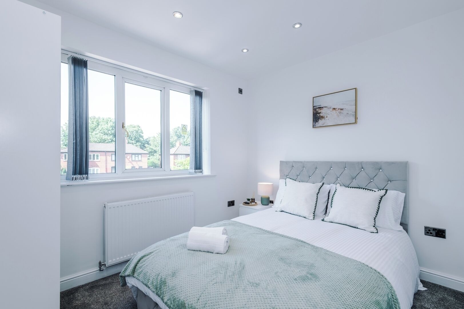 Une chambre à coucher avec un lit, une fenêtre, un radiateur, une table de chevet et un tableau accroché au mur.