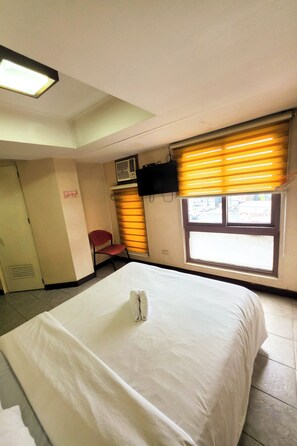A hotel room with a bed, a chair, a window with blinds, a television, and a door. - MGH Stays Quezon City (Quezon City)
