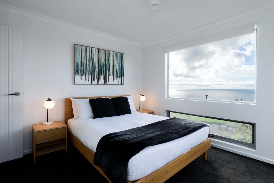 A modern bedroom with a large window, a bed with white bedding, two bedside tables with lamps, and a painting on the wall.