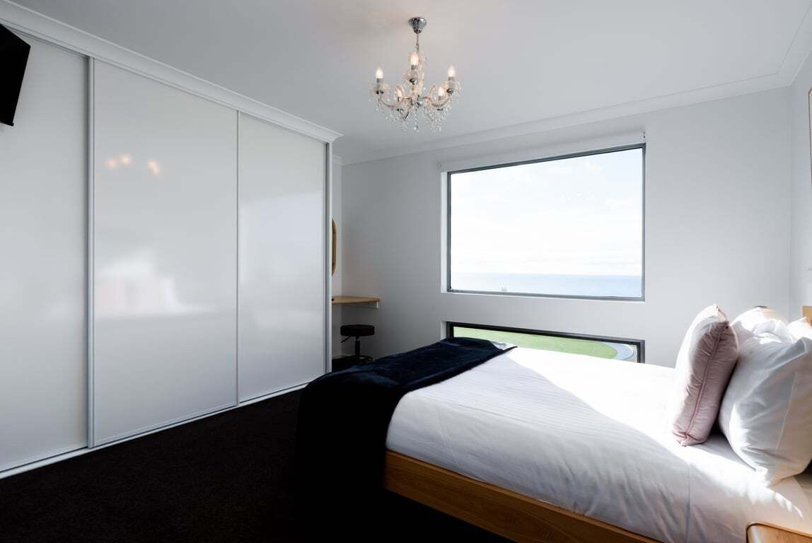 Un dormitorio moderno con un ventanal grande, una cama con almohadas, una mesita de noche y una araña.