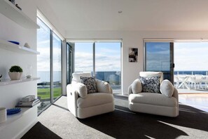 A modern living room with two white sofas, a bookshelf, and a large window offering a view of the sea.