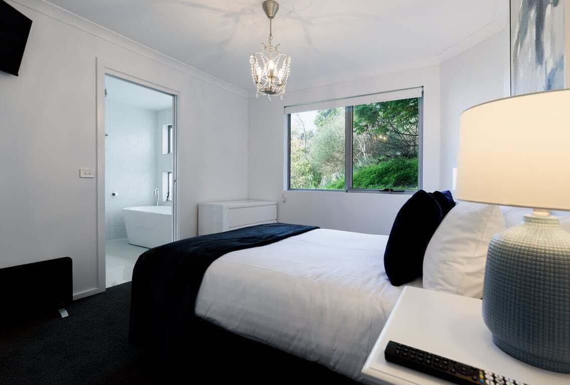 A bedroom with a bed, a nightstand, a lamp, a window with a view of greenery, and a door leading to a bathroom.