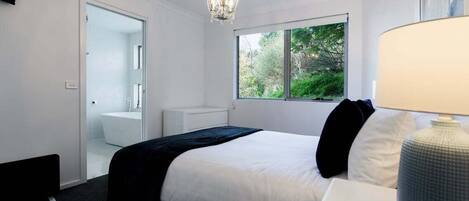 A bedroom with a bed, a nightstand, a lamp, a window with a view of greenery, and a door leading to a bathroom.