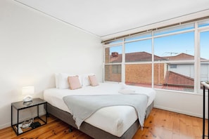 A bedroom with a large bed, a bedside table, and a window with a view of buildings. - Domi Rentals - The Murra Apartment (Murrumbeena)