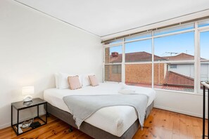 A bedroom with a large window, a bed with pillows, a bedside table with a lamp, and a wooden floor. - Domi Rentals - The Murra Apartment (Murrumbeena)