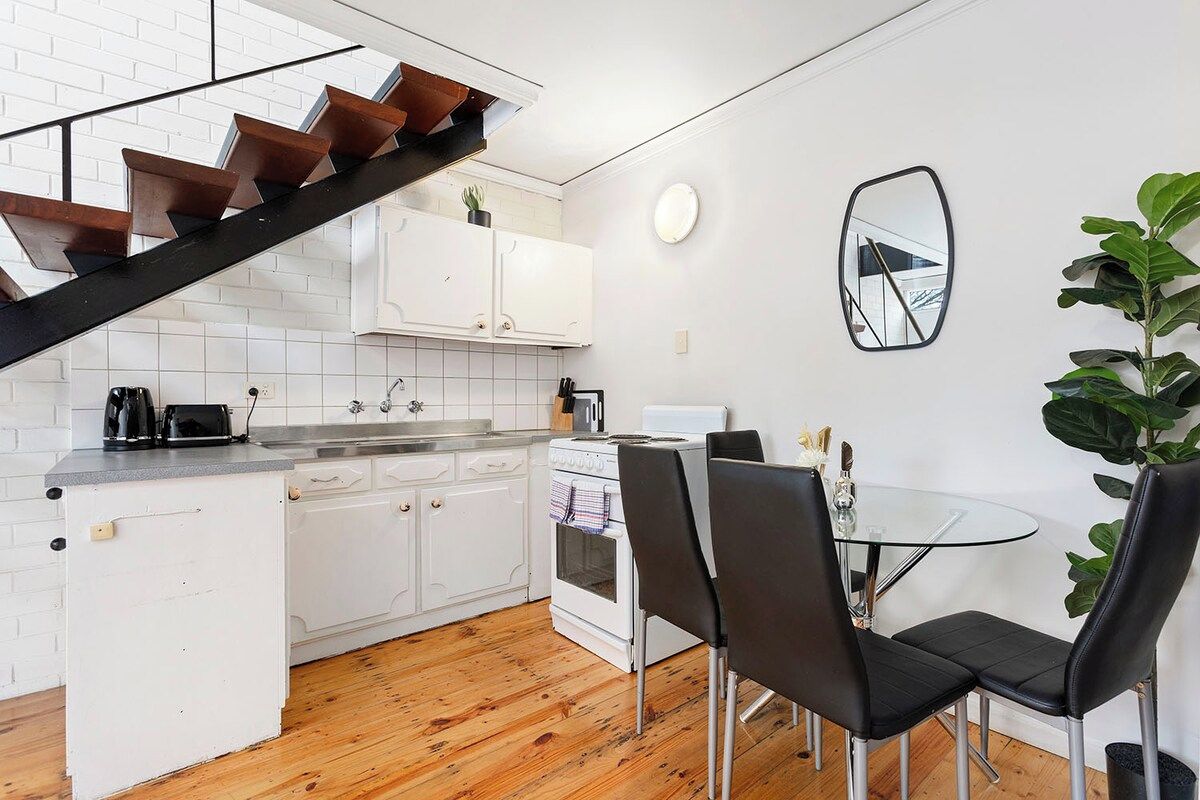 Una cocina moderna con armarios blancos, piso de madera, una zona de comedor con sillas negras y una escalera con escalones de madera.