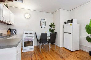 A compact kitchen with a stove, oven, microwave, and refrigerator. There is a dining table with chairs and a potted plant. - Domi Rentals - The Murra Apartment (Murrumbeena)