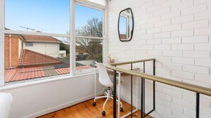 Une pièce avec un mur en briques blanches, une fenêtre donnant sur les maisons voisines, un fauteuil de bureau blanc et un miroir à cadre noir.