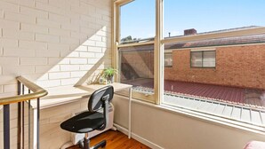 Une pièce avec une fenêtre, une chaise, une petite table avec une plante et une vue sur un bâtiment en briques à l’extérieur.