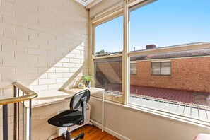 A room with a window, a chair, a small table with a plant, and a view of a brick building outside. - Domi Rentals - The Murra Apartments (Murrumbeena)
